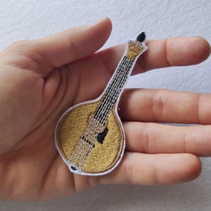 Pode incluir: Patch bordado de uma guitarra portuguesa, com um corpo dourado e um braço detalhado com costuras pretas e prateadas. O patch tem uma borda branca e é projetado para ser passado a ferro ou costurado no tecido.