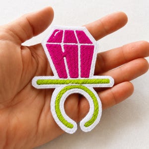 May include: An embroidered patch shaped like a ring pop candy. The top portion is a faceted magenta gemstone shape with white borders. The bottom portion is a lime green ring band with a white outline. The design is flat and stitched with thread.