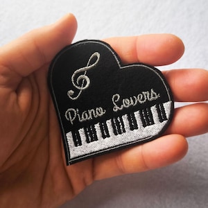 Pode incluir: Um emblema bordado em forma de coração com fundo preto, uma clave de sol prateada, uma fila de teclas de piano pretas e prateadas na parte inferior e o texto Piano Lovers bordado em fio prateado no centro do emblema.