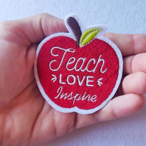 Könnte beinhalten: Gestickter Apfelaufnäher mit den Worten "Teach Love Inspire" in weißer Schrift. Der Apfel ist rot mit einem grünen Blatt und braunem Stiel, umrandet in Weiß. Ein charmantes Accessoire für Lehrer.