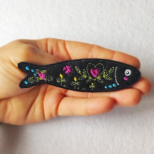 Può includere: Toppa ricamata nera a forma di pesce con un cuore e un design floreale in rosa, giallo e verde. Il pesce ha un occhio bianco e dettagli blu. La toppa è tenuta in una mano.