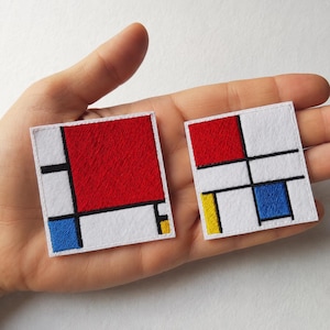 May include: Two square embroidered patches featuring a geometric design with white backgrounds, bold black grid lines, and blocks of primary colours including red, blue, and yellow. The patches have a clean, stitched border and a flat, fabric texture.