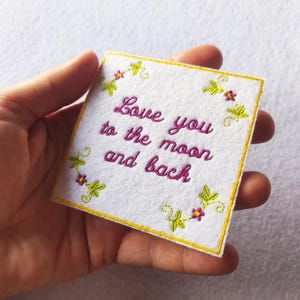 Peut inclure: Un patch carré en feutre blanc avec la phrase brodée en rose "Love you to the moon and back". Le patch est bordé d'une couture jaune et décoré d'éléments floraux verts et violets.