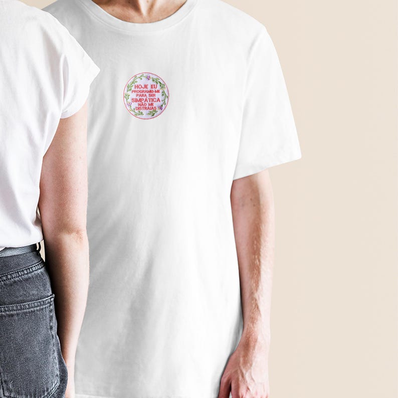 以下が含まれることがあります： 胸に円形のフローラルデザインが施された白いTシャツ。デザインは、緑の葉とピンクの花に囲まれたスクリプトフォントのテキストが特徴です。シャツは柔らかく、軽量な素材で作られています。