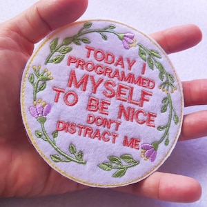 Puede incluir: Un parche bordado redondo y blanco con el texto rojo "TODAY I PROGRAMMED MYSELF TO BE NICE DON'T DISTRACT ME". El parche está rodeado por un diseño floral verde con flores moradas.