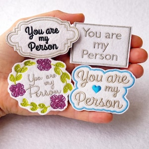 Puede incluir: Cuatro parches bordados con la frase "You are my Person". Los diseños incluyen un rectángulo blanco, una corona floral y dos formas decorativas con acentos azules, verdes y morados. El texto está en cursiva.