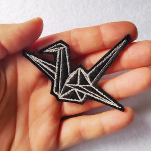 Può includere: Toppa a forma di gru in feltro nero con dettagli ricamati bianchi. L'uccello in stile origami presenta un design geometrico con un contorno dettagliato. La toppa è tenuta in mano, mostrando le dimensioni e la consistenza dell'oggetto artigianale.