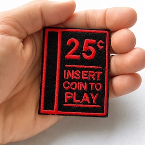 Könnte beinhalten: Ein rechteckiger, schwarz bestickter Aufnäher mit rotem Rand und rotem Text, der 25 cents, INSERT COIN TO PLAY lautet. Das Design zeigt einen vertikalen roten Streifen auf der linken Seite und horizontale rote Linien zwischen den Textzeilen.