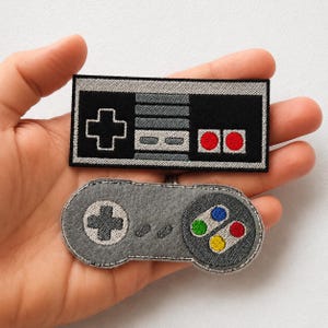 Puede incluir: Dos parches bordados con forma de mandos de videojuegos retro. El parche rectangular superior es negro, gris y blanco con dos botones rojos. El parche ovalado inferior es gris con una cruceta negra y botones de colores azul, verde, rojo y amarillo.