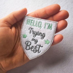 Peut inclure: Un écusson gris en forme de bouclier avec le texte brodé "HELLO, I'M Trying my Best" en vert et noir. Deux étoiles vertes ornent le texte. L'écusson est tenu dans une main.