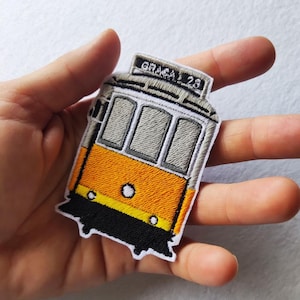 Puede incluir: Parche bordado que representa un tranvía amarillo y gris de Lisboa, con el texto "GRACA 28". El parche tiene un borde negro y está diseñado para planchar o coser sobre tela. Un artículo de recuerdo o con temática de viaje.