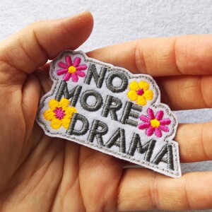 Peut inclure: Un écusson brodé blanc avec les mots "NO MORE DRAMA" en lettres grises. L'écusson est orné de fleurs brodées jaunes et roses et d'une bordure grise. L'écusson est tenu dans une main.