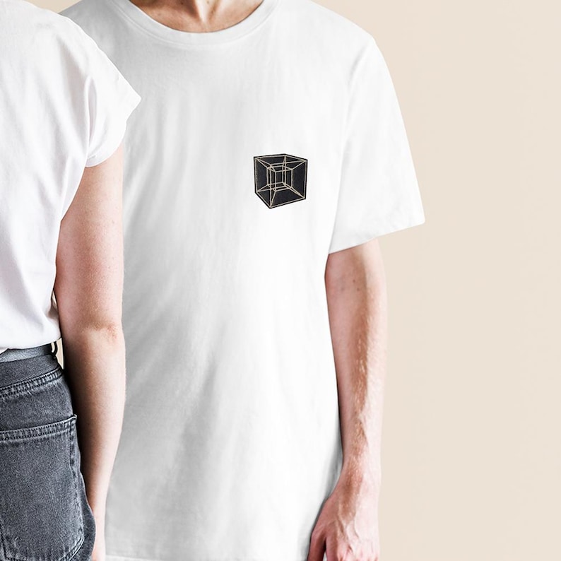 May include: White t-shirt with a small, geometric cube design on the chest. The cube is black with gold-colored outlines, creating a minimalist and modern aesthetic. The shirt is a classic crew neck style.