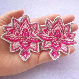 Könnte beinhalten: Zwei gestickte Lotusblüten-Patches. Die Patches sind in leuchtendem Pink gehalten und weisen detaillierte Stickereien auf, die mit hellem Beige und Weiß umrandet sind. Das Lotusblüten-Design ist ein beliebtes Motiv in Kunsthandwerk und Mode.