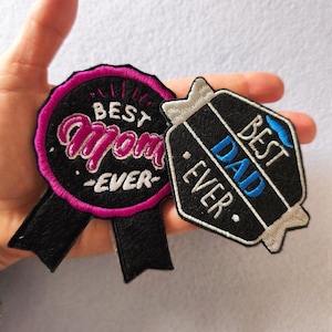 May include: Two embroidered patches, one round and one hexagonal. The round patch says "Best Mom Ever" in pink and white on a black background with a pink border. The hexagonal patch says "Best Dad Ever" in blue and white on a black background.
