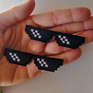 Könnte beinhalten: Zwei schwarze gestickte Aufnäher in Form einer verpixelten Sonnenbrille. Jeder Aufnäher hat einen rechteckigen Rahmen mit gezacktem Rand und weißen gestickten Quadraten als Gläser, was eine markante digitale 8-Bit-Optik erzeugt.
