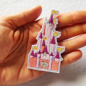 Pode incluir: Patch bordado com um castelo rosa com torres roxas e bandeiras amarelas. O castelo tem um design em forma de coração na base e é delineado com uma borda cinza. O patch é feito de tecido e tem um design costurado.