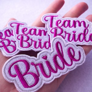 May include: Embroidered patches with the words "Team Bride" and "Bride" in a cursive font. The patches have a white felt base with a silver border and bright pink lettering.