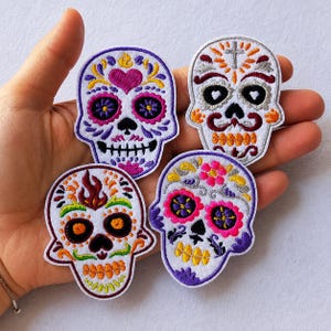Könnte beinhalten: Vier bestickte Totenkopf-Aufnäher mit komplexen, farbenfrohen Designs. Jeder Aufnäher zeigt einzigartige Muster in Lila, Pink, Orange, Gelb und Grün auf weißem Grund, mit detaillierten Stickereien und dekorativen floralen Elementen.