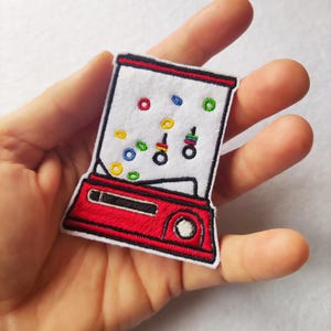 Könnte beinhalten: Ein gestickter Aufnäher in Form eines Kaugummiautomaten. Der Aufnäher hat eine rote Basis und einen weißen oberen Teil mit bunten Gumballs. Schwarze Details umreißen das Design. Der Aufnäher wird in einer Hand gehalten.