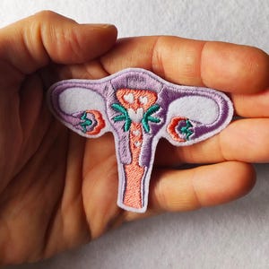 May include: Embroidered patch depicting a stylized human uterus. The patch features a purple outline with white, peach, and teal accents. The design includes details of the fallopian tubes and ovaries, with a light pink background.