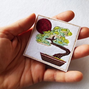 Pode incluir: Patch bordado com uma árvore bonsai e um sol vermelho ao fundo. A árvore tem folhagem verde e azul clara, um tronco castanho e está num vaso castanho. O patch é quadrado com uma borda dourada.