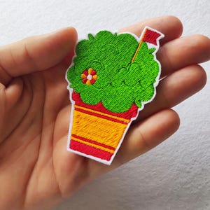 Pode incluir: Patch bordado com um design verde e fofo que se assemelha a uma bebida, com um copo vermelho e amarelo. Uma pequena flor e uma palhinha com uma bandeira vermelha também são visíveis. O patch é delineado em branco.