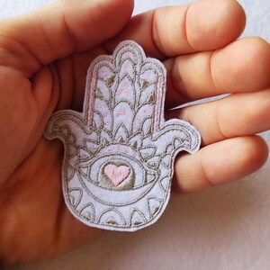 Peut inclure: Un écusson en forme de main de Hamsa violet clair avec des broderies complexes marron clair et roses. Le patch présente un cœur au centre et est tenu dans une main.