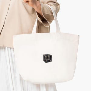 May include: Off-white canvas tote bag with a geometric cube design in black and gold. The bag has sturdy white handles and is held by a person wearing a beige jacket and white skirt.