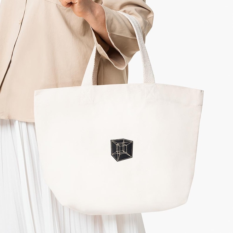 May include: Off-white canvas tote bag with a geometric cube design in black and gold. The bag has sturdy white handles and is held by a person wearing a beige jacket and white skirt.