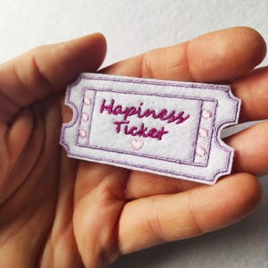 Könnte beinhalten: Ein weißes Filz-Ticket mit den Worten "Happiness Ticket" in Pink gestickt. Das Ticket hat einen lila Rand und kleine Herz-Akzente. Das Ticket wird in einer Hand gehalten.