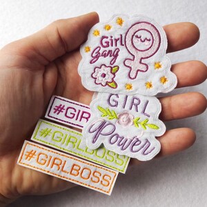 Pode incluir: Uma coleção de patches bordados com temas feministas. Os patches apresentam frases como "Girl Gang", "Girl Power" e "Girl Boss", juntamente com um símbolo feminino e desenhos florais. Os patches são em várias cores, incluindo branco, roxo, verde e laranja.