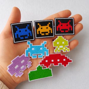 Pode incluir: Uma coleção de emblemas bordados com designs pixelizados de alienígenas e naves espaciais em várias cores, incluindo azul, amarelo, vermelho, azul claro, verde limão, roxo e verde. Os emblemas são quadrados ou moldados como personagens retro.