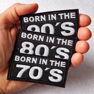 Könnte beinhalten: Drei schwarze, rechteckige Aufnäher mit weißem, gesticktem Text. Die Aufnäher lesen "BORN IN THE 90'S", "BORN IN THE 80'S" und "BORN IN THE 70'S". Die Aufnäher sind übereinander gestapelt.
