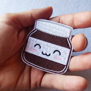 Pode incluir: Patch bordado com um pote sorridente de creme de avelã e chocolate. O patch tem um design marrom e branco com um rosto fofo e bochechas rosadas. O patch é contornado em marrom e tem um topo branco.