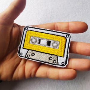 May include: Embroidered patch in the shape of a yellow cassette tape with a white border. The cassette tape has gray and black details, including the tape reels and the tape window. The patch is held in a person's hand.