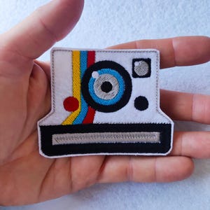 Pode incluir: Um patch de feltro branco em forma de câmera instantânea vintage. A câmera tem uma lente azul grande, uma borda preta e listras coloridas amarelas, azuis e vermelhas. Um pequeno círculo vermelho e um ponto preto também são visíveis.