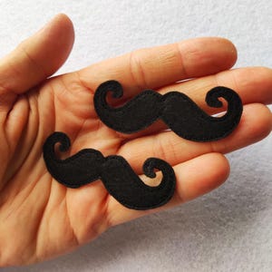 May include: Two black felt mustache-shaped appliques with stitched edges. Each piece features a classic handlebar design with curled ends, crafted from a soft, matte fabric material suitable for various decorative craft projects and embellishments.