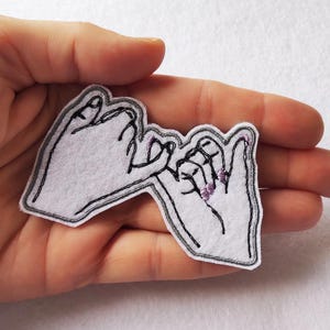 Peut inclure: Un patch en feutre blanc représentant un dessin à la main de deux mains faisant un serment de doigts. Le patch a des contours brodés noirs et une bordure grise. Les ongles sont peints en violet.