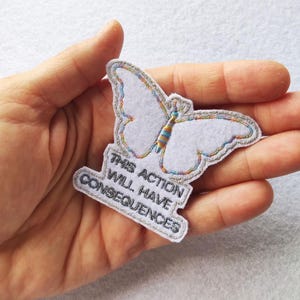 Puede incluir: Un parche de fieltro blanco con forma de mariposa con un contorno plateado y detalles coloridos. El parche tiene el texto "THIS ACTION WILL HAVE CONSEQUENCES" en un rectángulo.