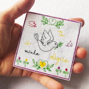 Pode incluir: Bordado quadrado em feltro branco com uma pomba carregando um ramo de oliveira, rodeado por texto bordado e desenhos florais em verde, amarelo e vermelho. As bordas são aparadas com uma borda roxa.