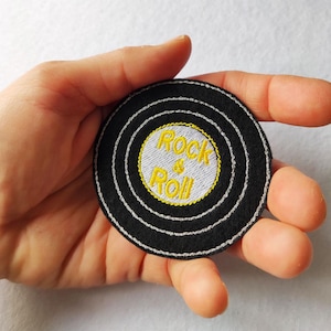 Puede incluir: Un parche bordado circular con forma de disco de vinilo con fondo negro, anillos concéntricos plateados y un círculo central plateado que presenta el texto bordado en amarillo Rock & Roll. El diseño imita un disco de música clásico.