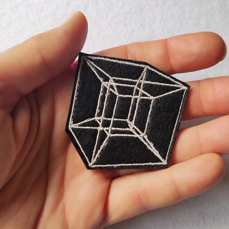 May include: Black geometric patch with a white embroidered 3D cube design. The patch is held in a hand, showcasing the intricate details of the stitched lines. The patch has a unique shape, adding to its visual appeal.