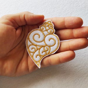 Pode incluir: Patch bordado em forma de coração branco e dourado. O patch apresenta uma base branca com bordados dourados intrincados em espiral. O design é um coração estilizado com um topo decorativo e uma parte inferior pontiaguda.