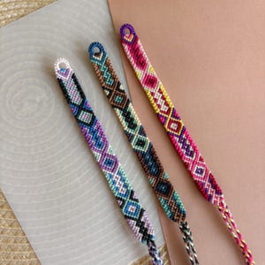 Peut inclure: Trois bracelets d'amitié tissés à la main présentant des motifs géométriques en losange complexes. Le premier bracelet utilise du fil violet, bleu et blanc. Le deuxième utilise du marron, du bleu sarcelle et du noir. Le troisième utilise du rose, du jaune et du blanc.