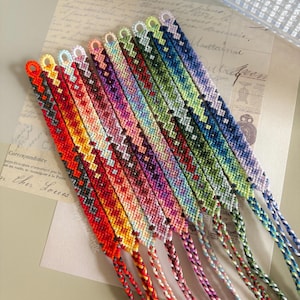 Peut inclure: Une collection de bracelets d'amitié colorés. Chaque bracelet présente un motif tissé dans une variété de couleurs, notamment le rouge, l'orange, le jaune, le rose, le violet, le vert et le bleu. Les bracelets sont exposés sur une surface claire.