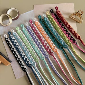 Peut inclure: Une collection de bracelets d'amitié colorés avec un motif géométrique. Chaque bracelet présente une palette de couleurs différente, notamment noir, bleu, vert, orange et rose. Les bracelets sont faits de fil tissé et ont une boucle en haut pour attacher.