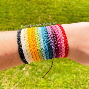 May include: A stack of colourful braided friendship bracelets in various colours, including black, white, yellow, green, blue, purple, and red. The bracelets are tied together with a brown string.