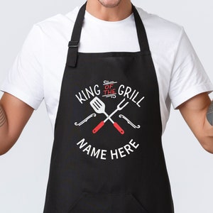 Pode incluir: Avental preto com o texto "KING OF THE GRILL" e "NAME HERE" em branco. O design inclui uma esp&aacute;tula e um garfo cruzados com cabos vermelhos. O avental tem uma al&ccedil;a de pesco&ccedil;o preta e &eacute; usado por uma pessoa vestindo uma camiseta branca.