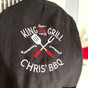 Pode incluir: Avental preto com texto bordado branco e um design de esp&aacute;tula e garfo cruzados. O texto diz "KING OF THE GRILL" acima de "CHRIS' BBQ". A esp&aacute;tula e o garfo t&ecirc;m cabos vermelhos. O avental est&aacute; pendurado.